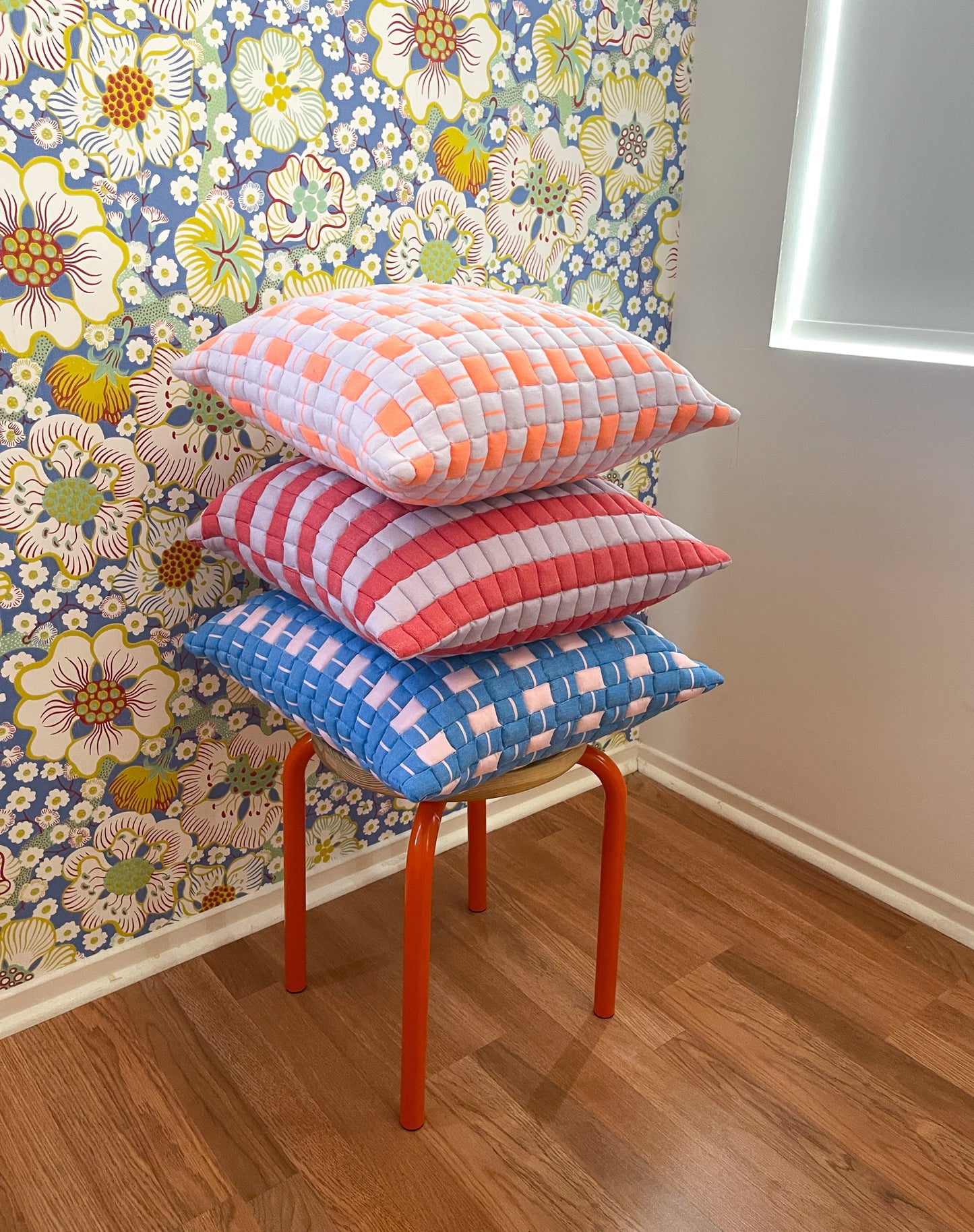 Stack of colorful checkered pillows on a stool against a floral wallpapered wall.