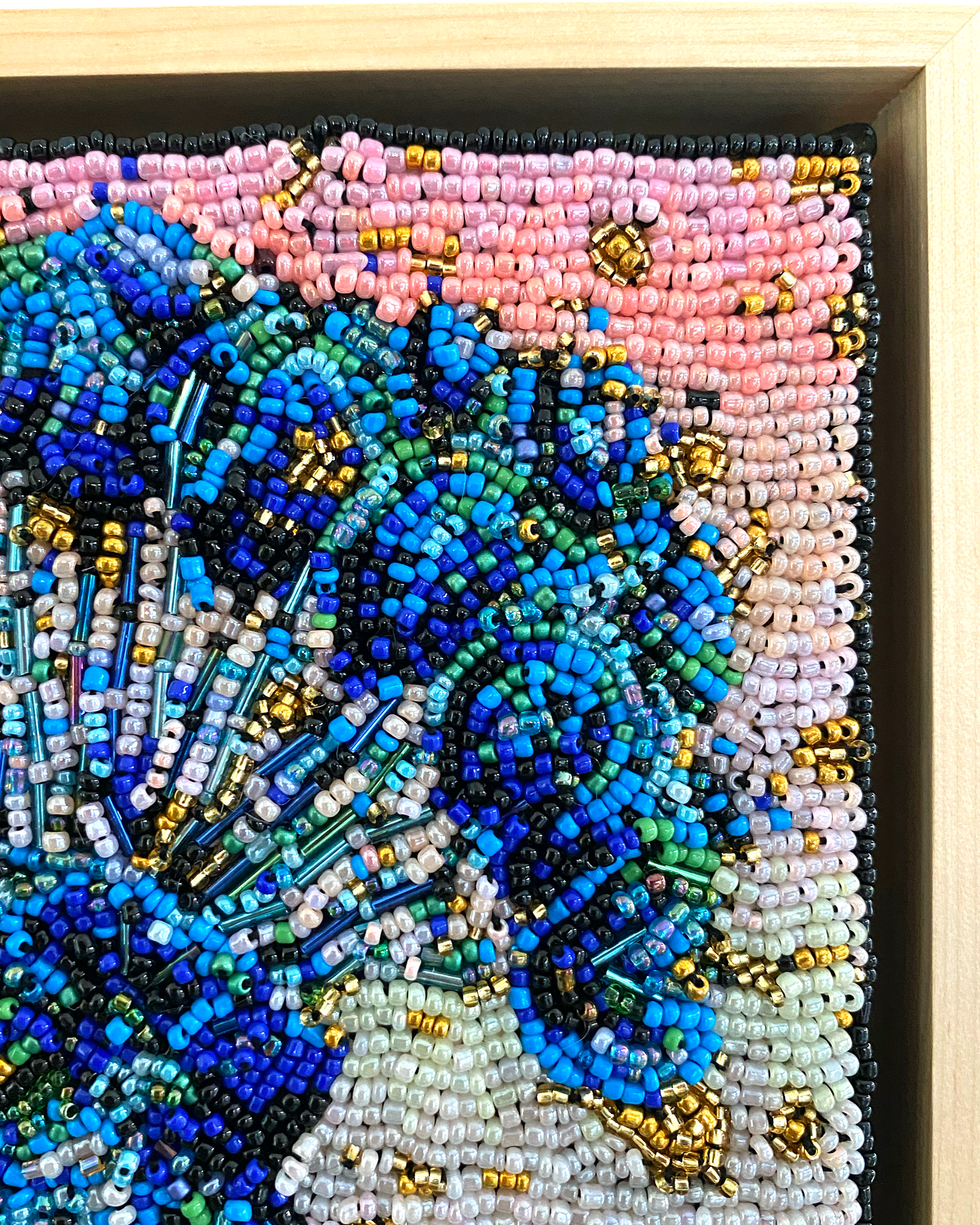 Close-up of a beaded artwork with blue and pink colors, framed in beige.