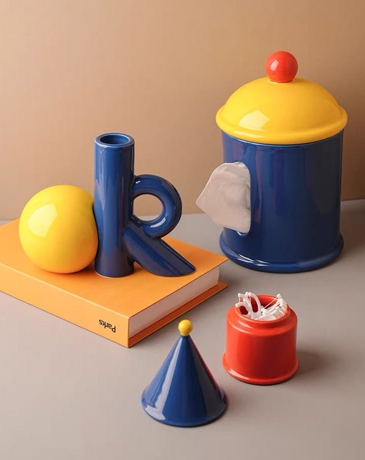 Colorful ceramic items including a blue vase, yellow ball, red container, and blue cone on a beige background.
